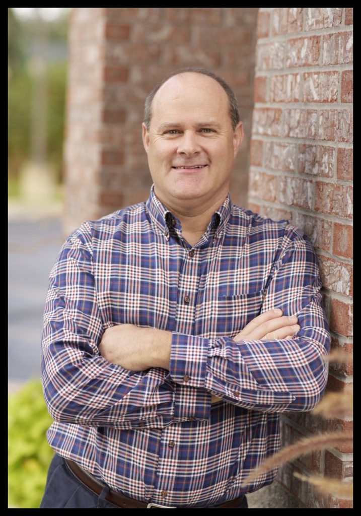 A doctor in a button up shirt casually leaning against a brick wall with his arms humbly crossed for a photo with a kind smile on his face.